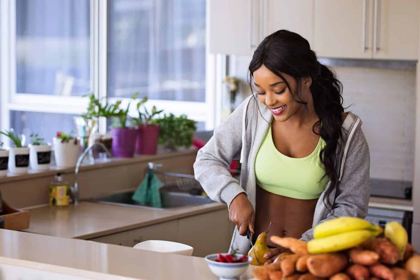 Get fit at home: smiling woman in a sports bra and grey tracksuit jacket cutting a pear in her kitchen with fresh fruit and vegetables in the foreground.