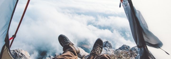 Frugal camping tips: a man's feet are visible from the opening of a tent, looking out over a landscape covered by clouds.