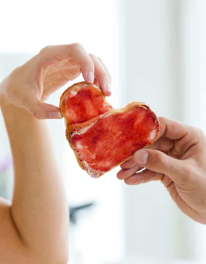 Two hands holding a heart-shaped piece of toast with red jam.