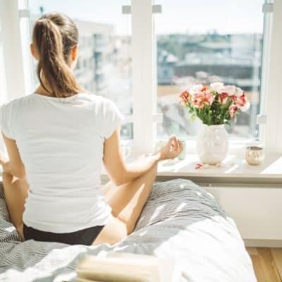 How to live a more intentional life: girl meditating on a bed overlooking a window