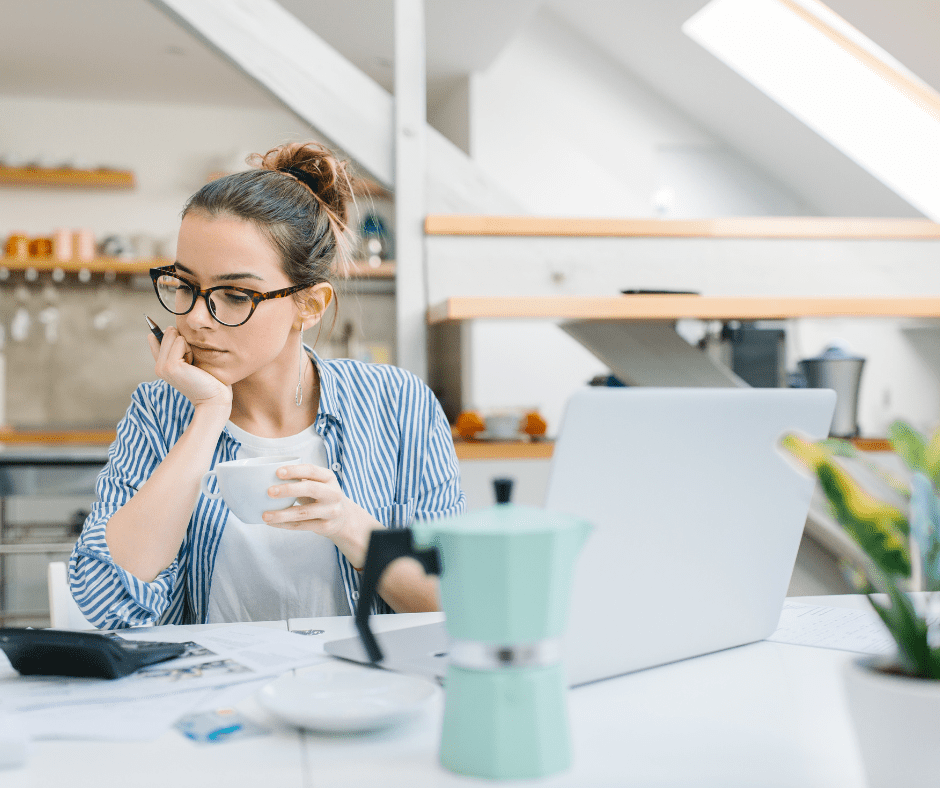 How to Budget When You're Paid Weekly: A Woman sits at a desk with an open laptop in front of her, holding a white mug, and looking towards a calculator that also sits on the desk.