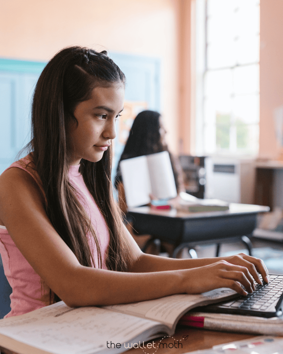 a girl sitting at a desk, typing on a keyboard. She has textbooks on the desk in front of her.